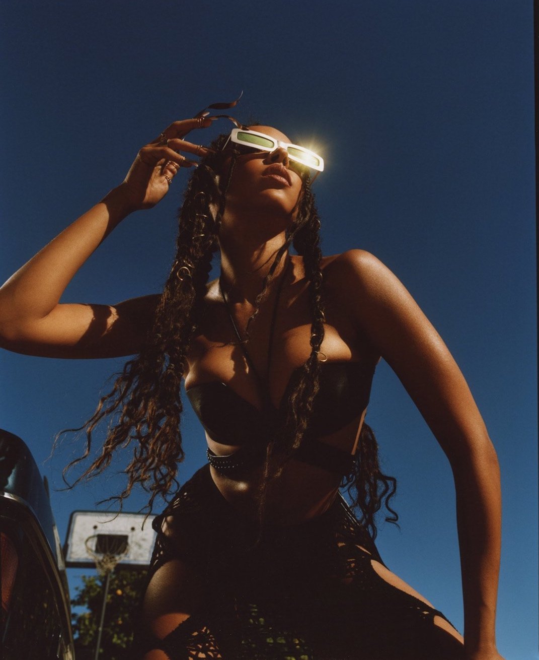 A dark-skinned woman poses in edgy fashion, wearing sunglasses, a black two-piece outfit with cutouts, and statement braids against a clear blue sky.