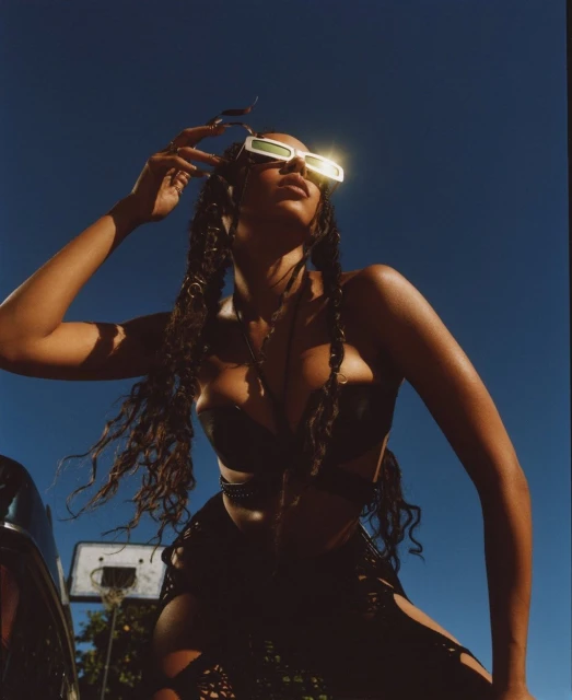 A dark-skinned woman poses in edgy fashion, wearing sunglasses, a black two-piece outfit with cutouts, and statement braids against a clear blue sky.