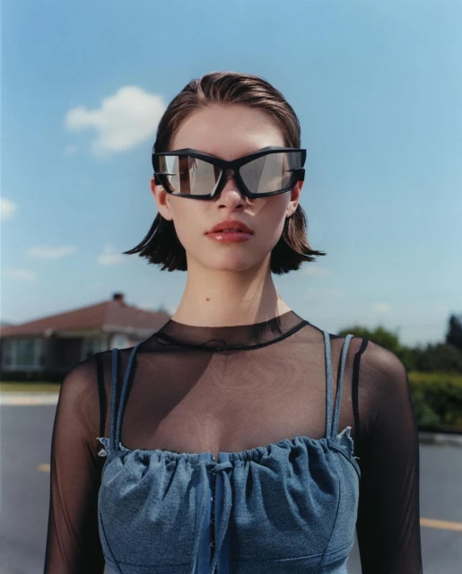 Portrait of a woman wearing oversized sunglasses, a sheer top and denim top, with a house and blue sky in the background.