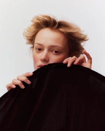 Close-up portrait of a woman with blonde hair holding a dark fabric garment covering the lower half of her face.