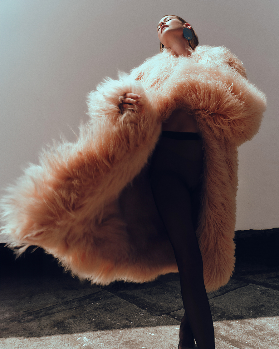 A woman poses in a large, fluffy pink fur coat and black tights against a plain backdrop, looking up with one earring visible.