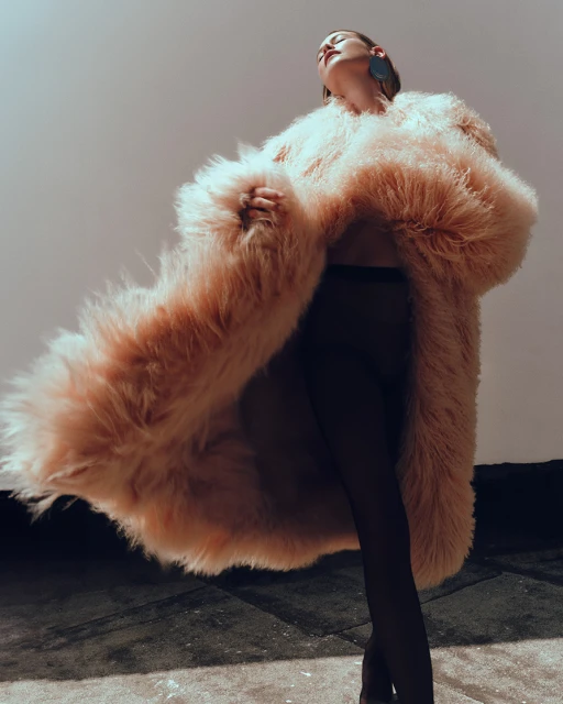 A woman poses in a large, fluffy pink fur coat and black tights against a plain backdrop, looking up with one earring visible.