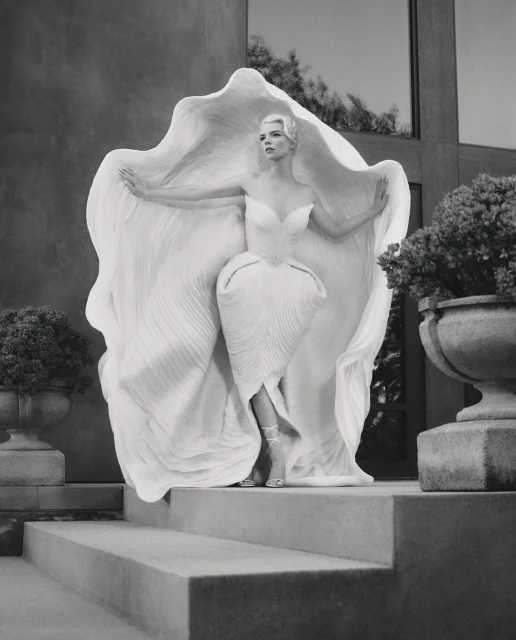 Monochromatic fashion portrait of a woman in an ornate white dress, posing on steps with potted plants and a building in the background.