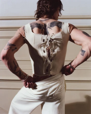 Shirtless male model with back tattoos poses in white pants and a lace-up top in a studio with neutral-toned wall paneling.