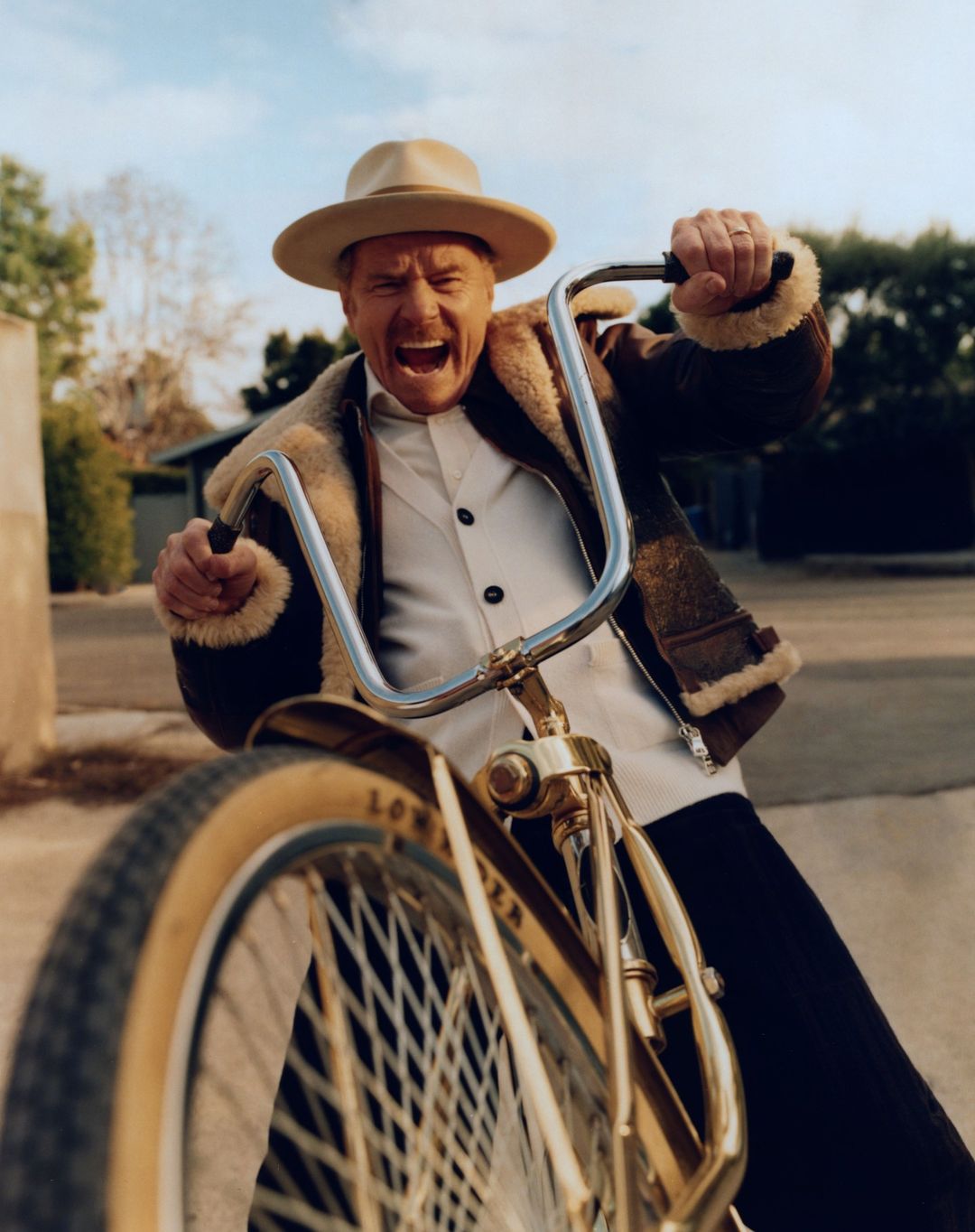 Excited mature man in a hat rides a vintage bicycle on a sunny street, shouting with joy and gripping the handlebars.