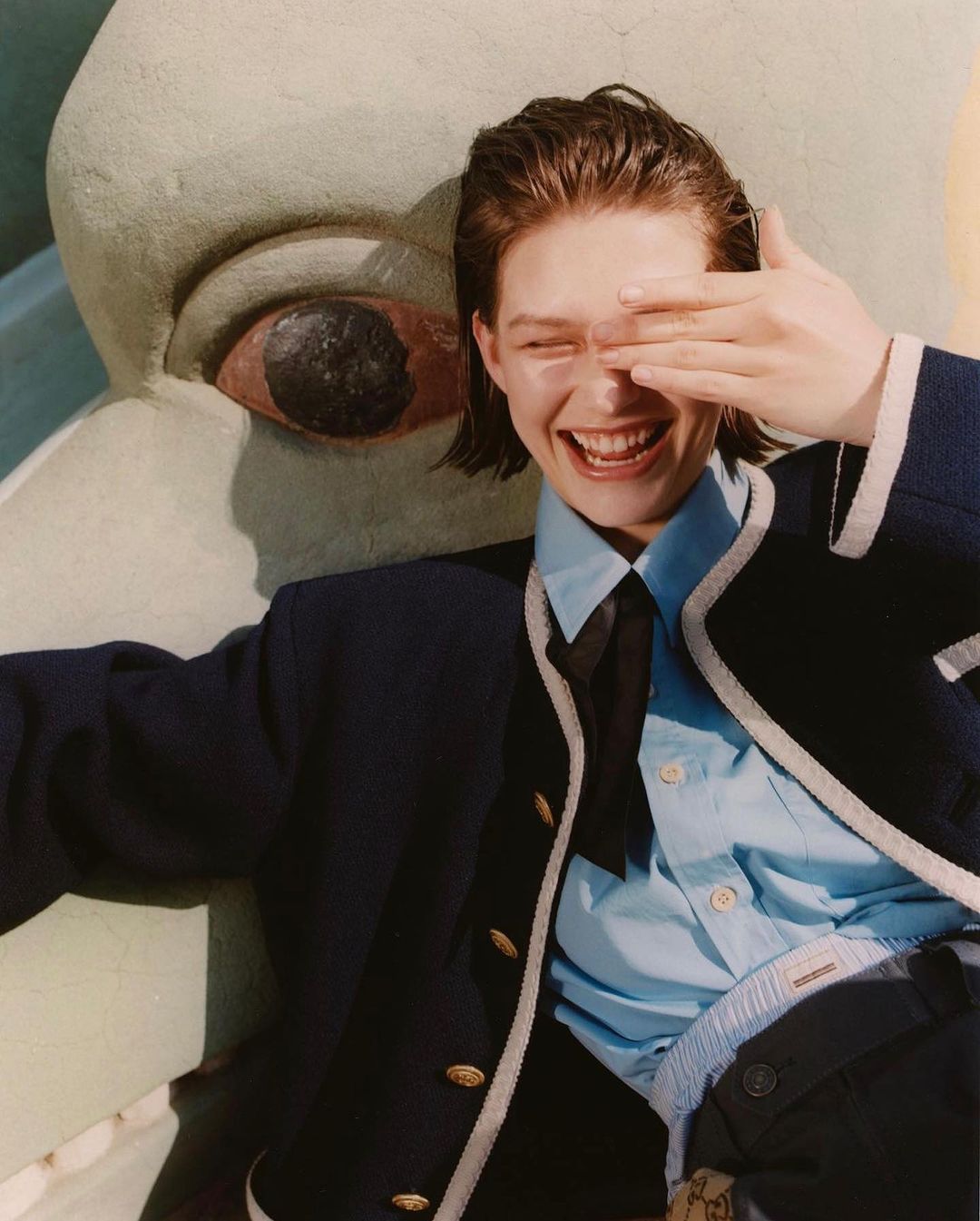 Smiling young adult woman wearing a blue blazer and shirt, covering one eye with her hand, posing outdoors near a painted sculpture.