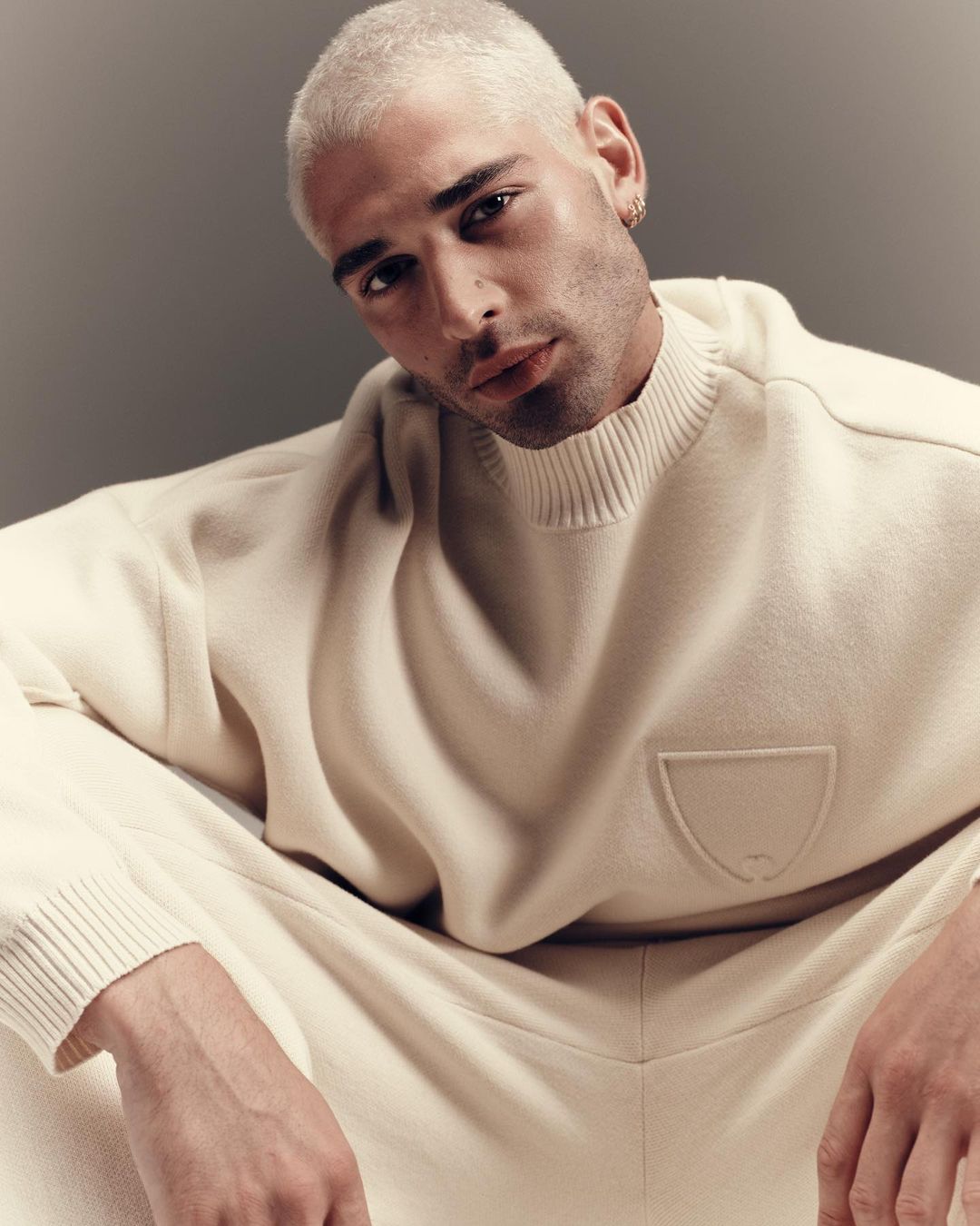 A young adult male with platinum blonde hair poses indoors, wearing a cream-colored mock neck sweater and matching sweatpants in a portrait.