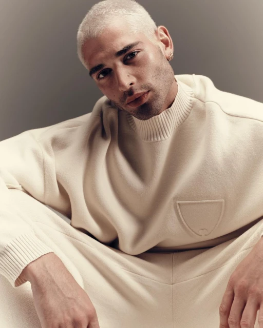 A young adult male with platinum blonde hair poses indoors, wearing a cream-colored mock neck sweater and matching sweatpants in a portrait.