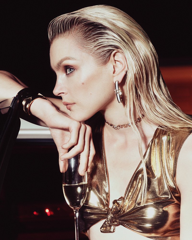 Elegant woman with blonde slicked-back hair and gold crop top leans on her arm with a glass of champagne, indoor setting with warm lighting.