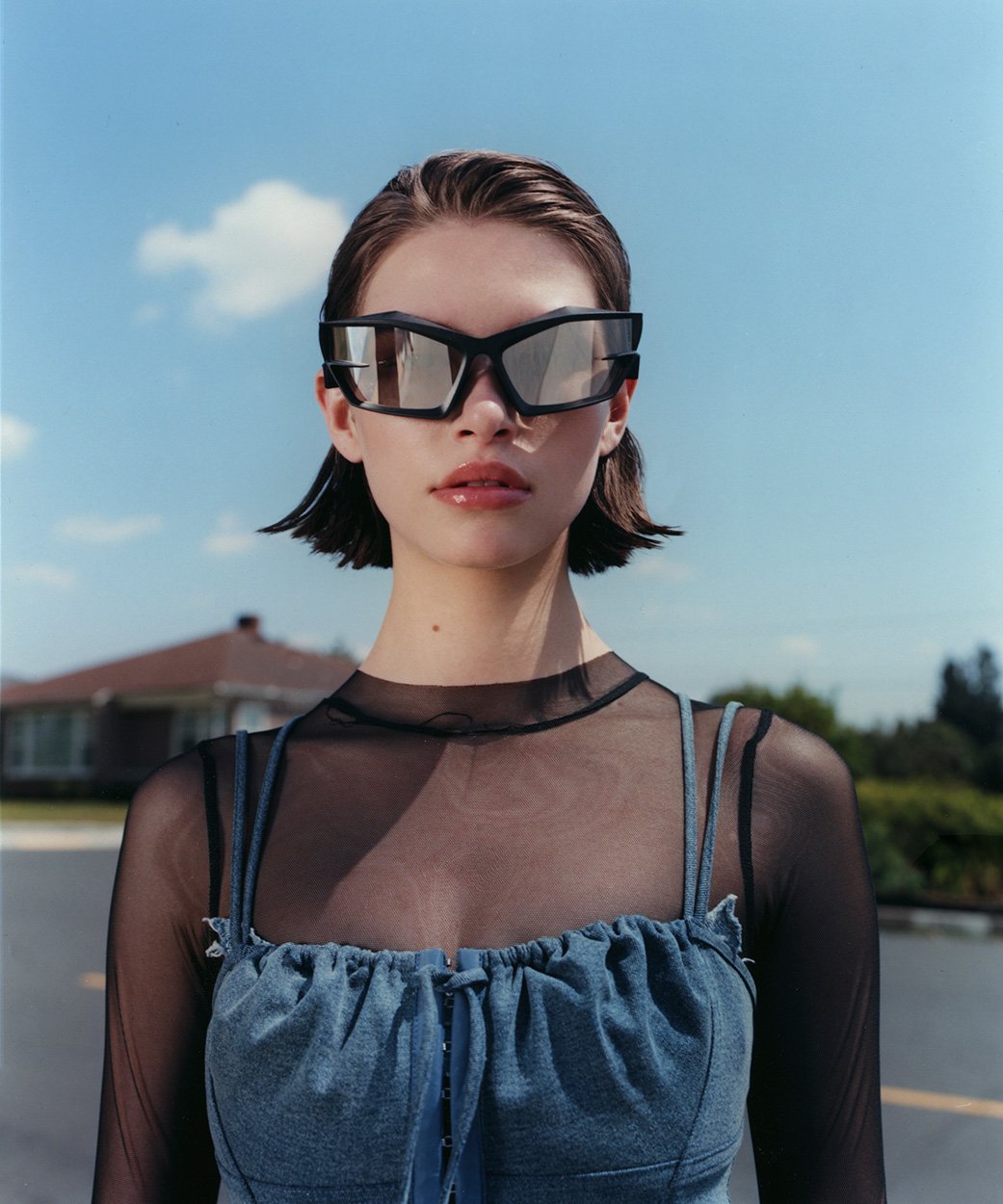 Woman with short hair wears futuristic sunglasses and denim top against a blue sky with clouds and residential background.