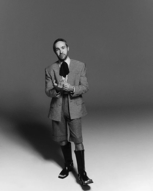 A full portrait of a man in a gray suit, black tie, and black boots, standing in a photo studio with a gray background and soft light.