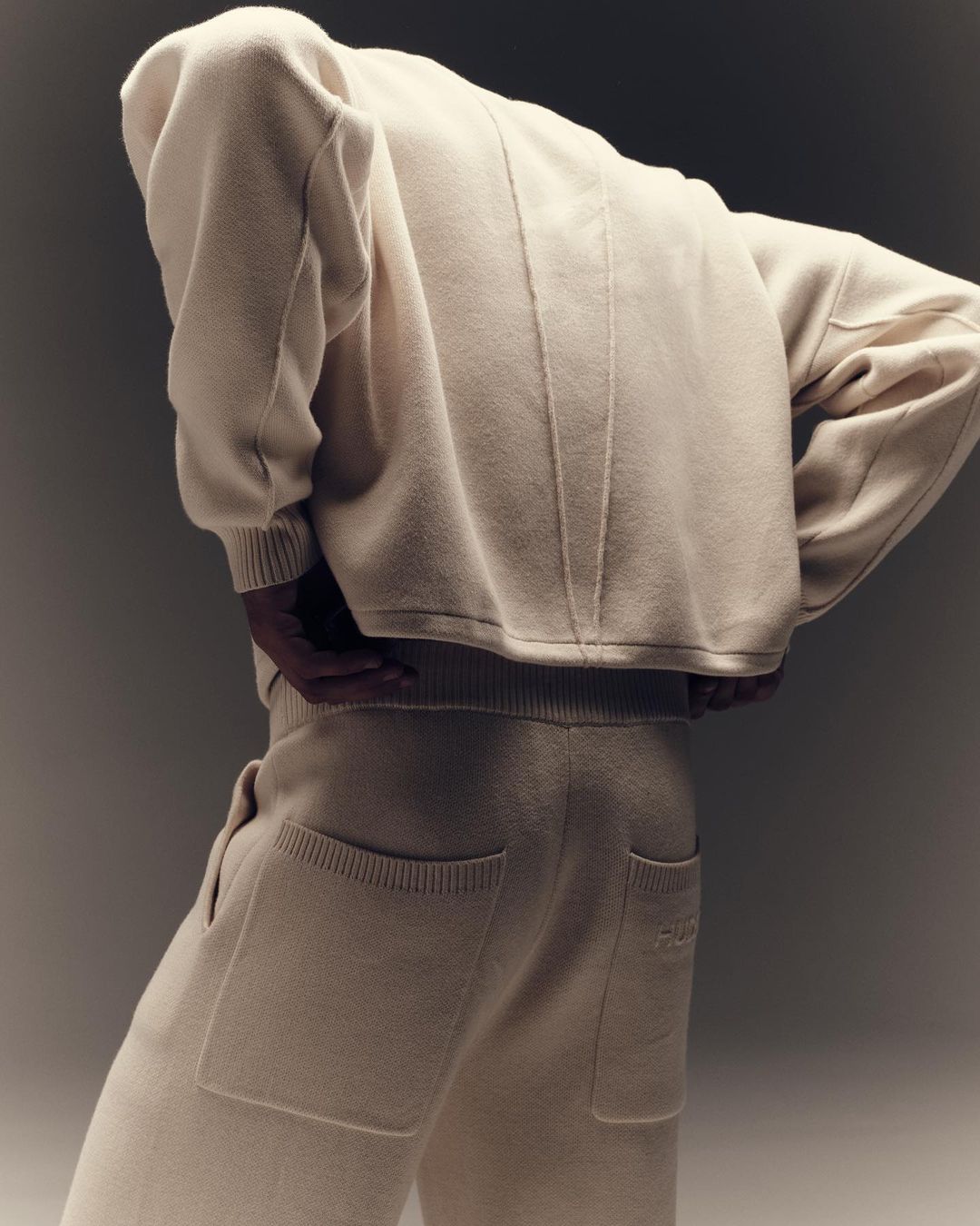 Person wearing a beige knitwear lounge set with a sweatshirt and sweatpants, posing in a studio with soft lighting and neutral background.