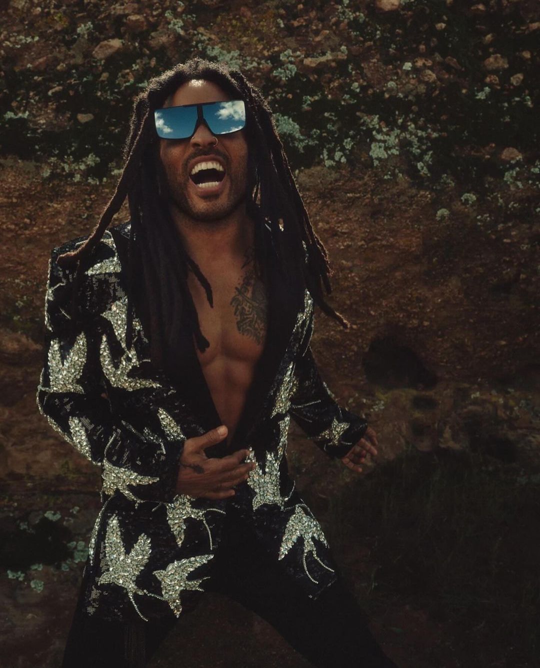 Adult male with dreadlocks, wearing sunglasses and a patterned jacket, poses against a rocky backdrop with an open mouth.