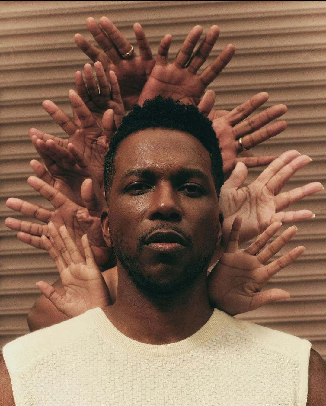 Portrait of a black man in a cream shirt, surrounded by a halo of hands against a tan ridged background. Studio lighting.