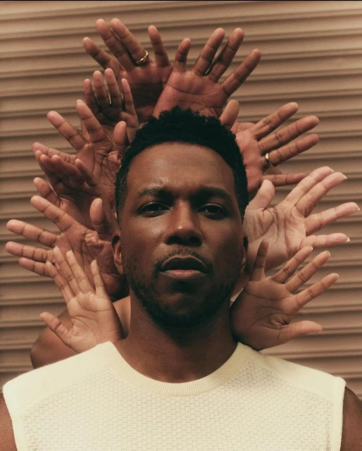 Portrait of a black man in a cream shirt, surrounded by a halo of hands against a tan ridged background. Studio lighting.