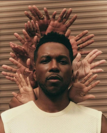 Portrait of a black man in a cream shirt, surrounded by a halo of hands against a tan ridged background. Studio lighting.