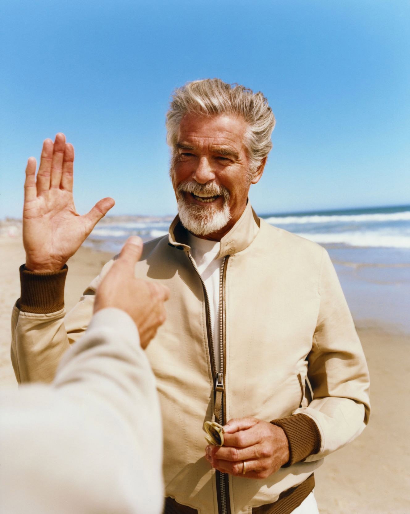 Smiling elderly man outdoors on a sandy beach raises his hand in greeting as another person points with the ocean and blue sky.