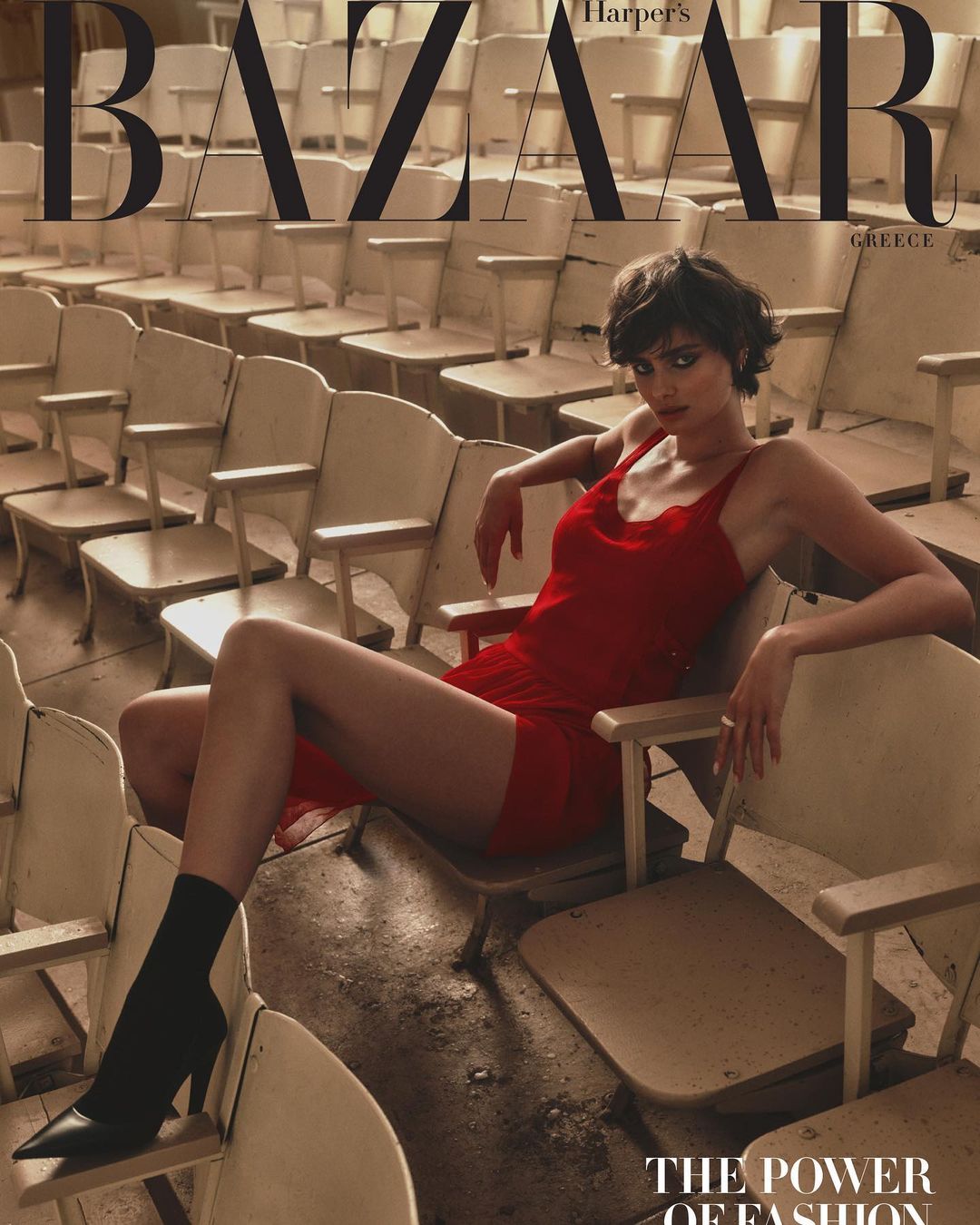 Fashion editorial featuring a woman in a red dress and black boots sitting among rows of theatre seats in a stark, vintage setting.