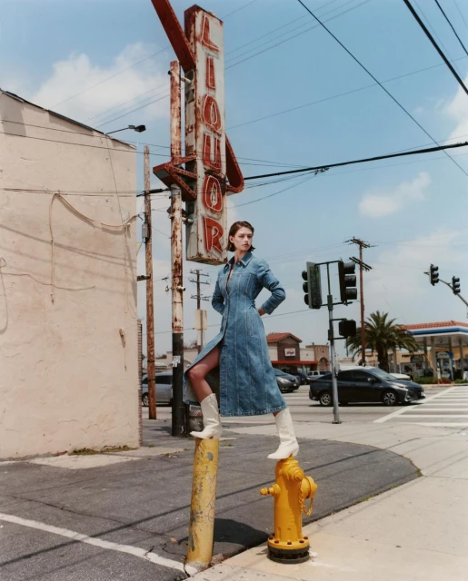 A woman poses on a yellow fire hydrant in a denim dress and white boots in an urban setting under a clear sky, with a liquor sign.