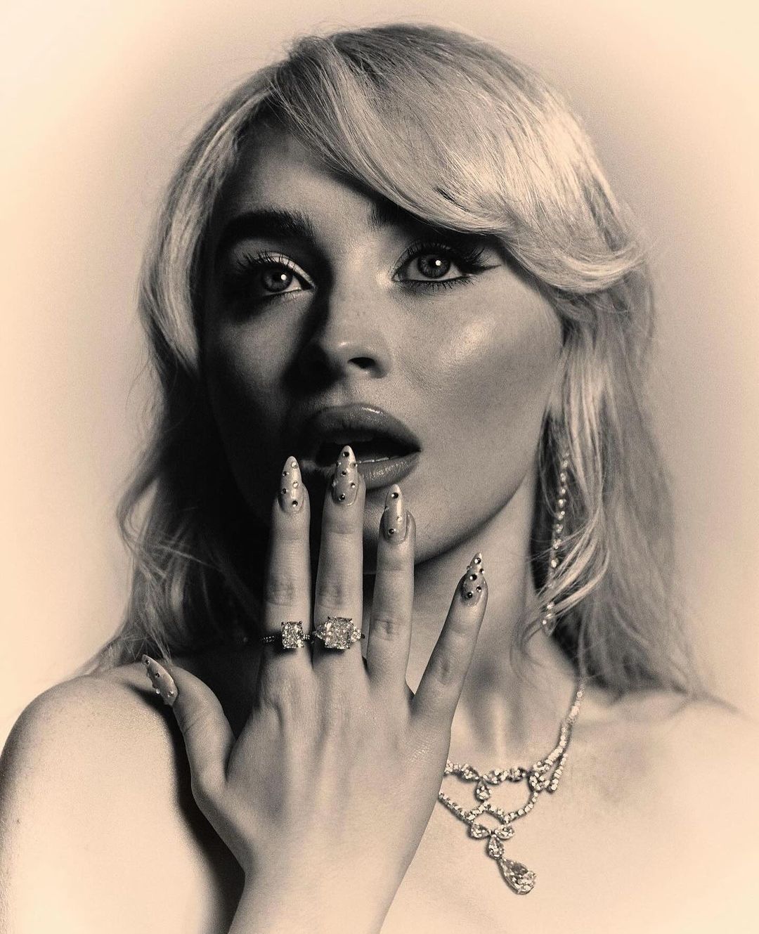 Monochrome portrait of a blonde woman wearing jeweled rings and necklace, hand near her face, against a neutral background.