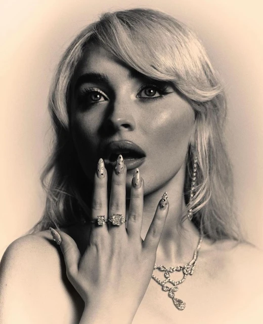 Monochrome portrait of a blonde woman wearing jeweled rings and necklace, hand near her face, against a neutral background.