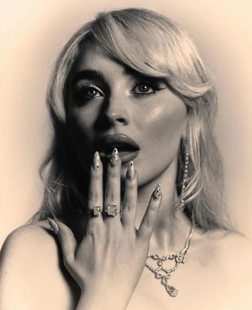 Monochrome portrait of a blonde woman wearing jeweled rings and necklace, hand near her face, against a neutral background.