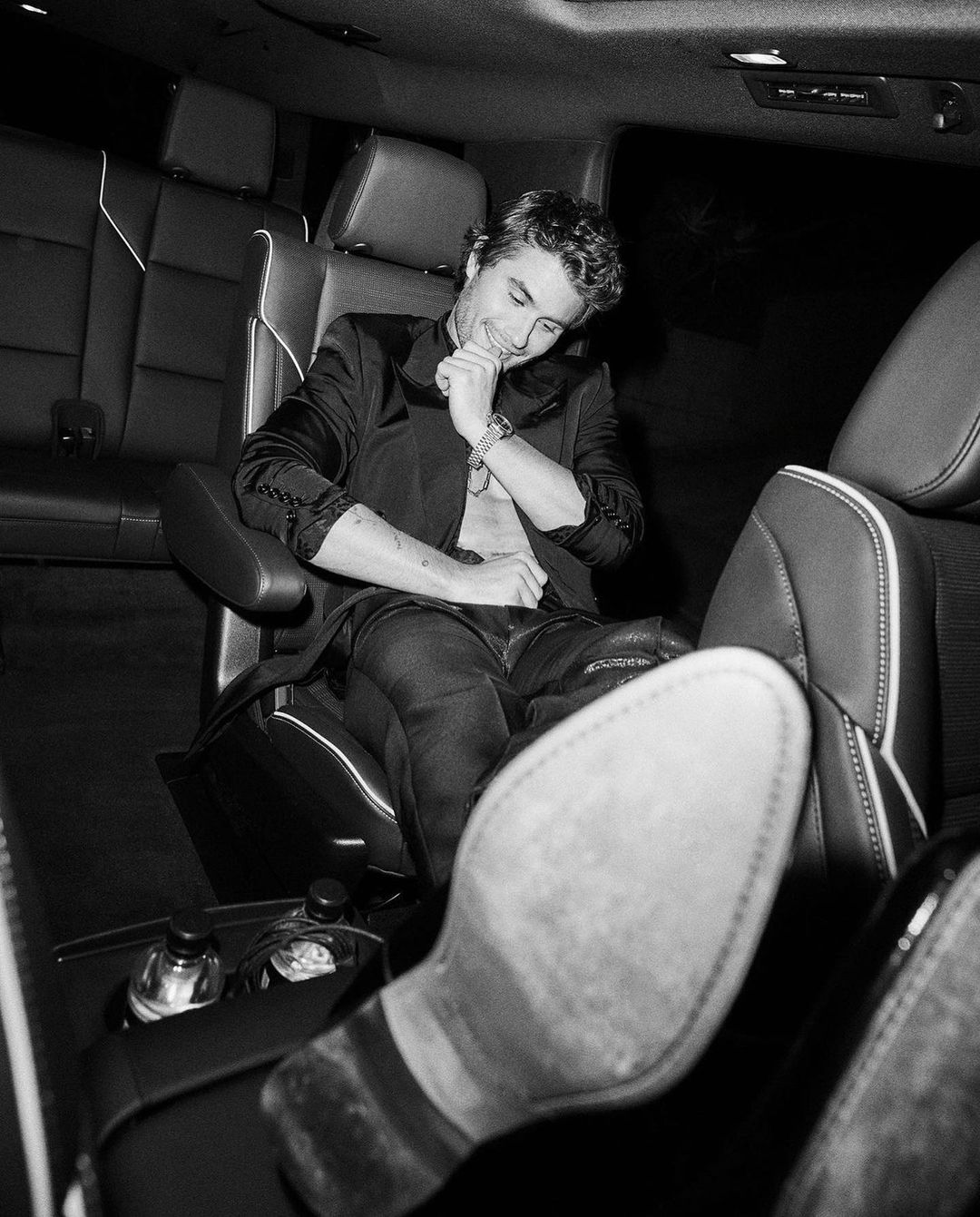 Monochromatic portrait of an adult male seated in a car, hand to face, wearing a blazer and jeans with his feet propped up.