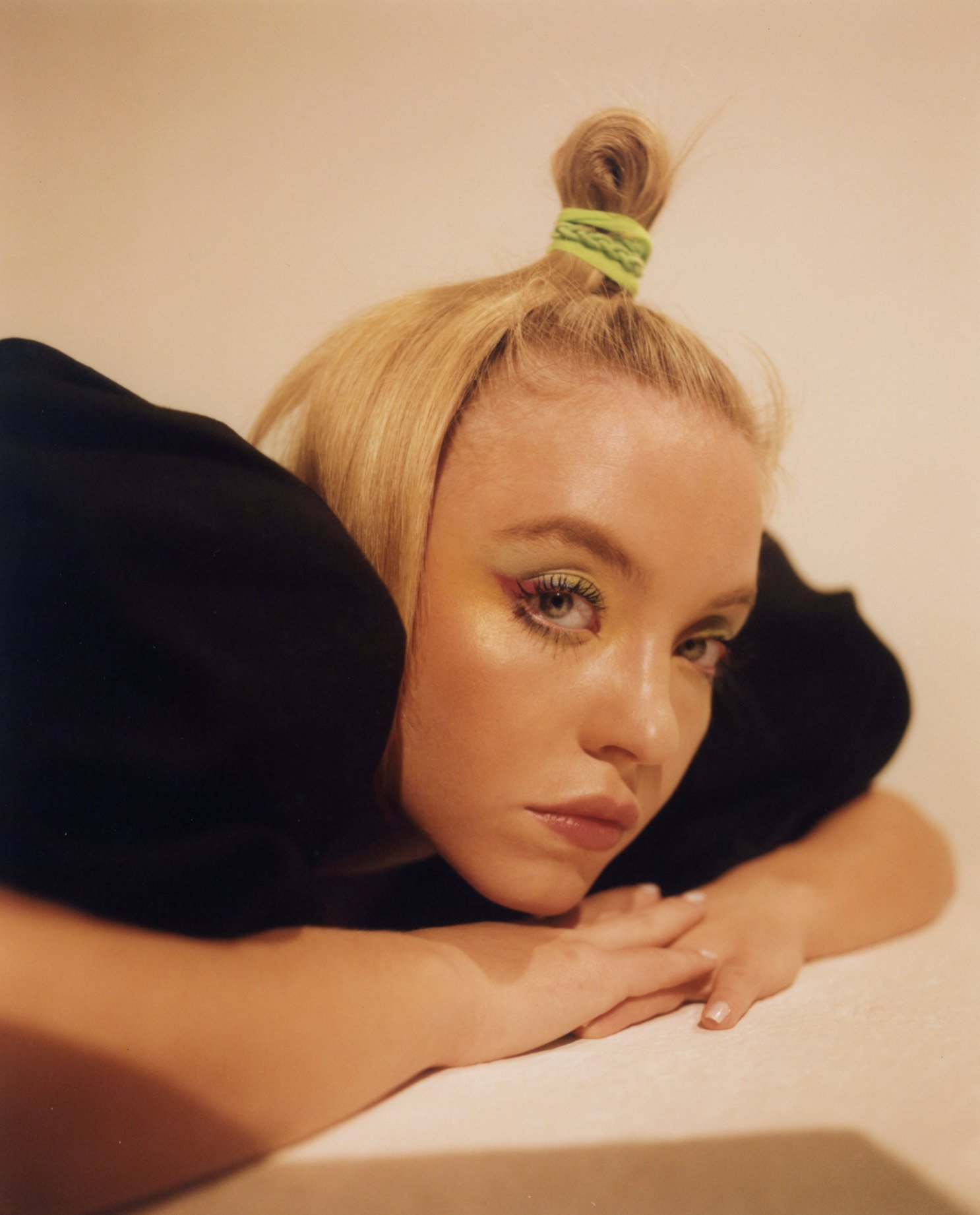 Woman with blond hair and colorful eyeshadow leans on her hands with a neutral expression in a studio with a tan and white backdrop.
