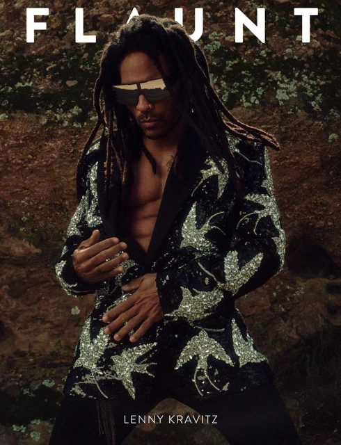 Lenny Kravitz poses on a rock, wearing sunglasses and a black blazer decorated with silver bird patterns for a magazine cover shoot.