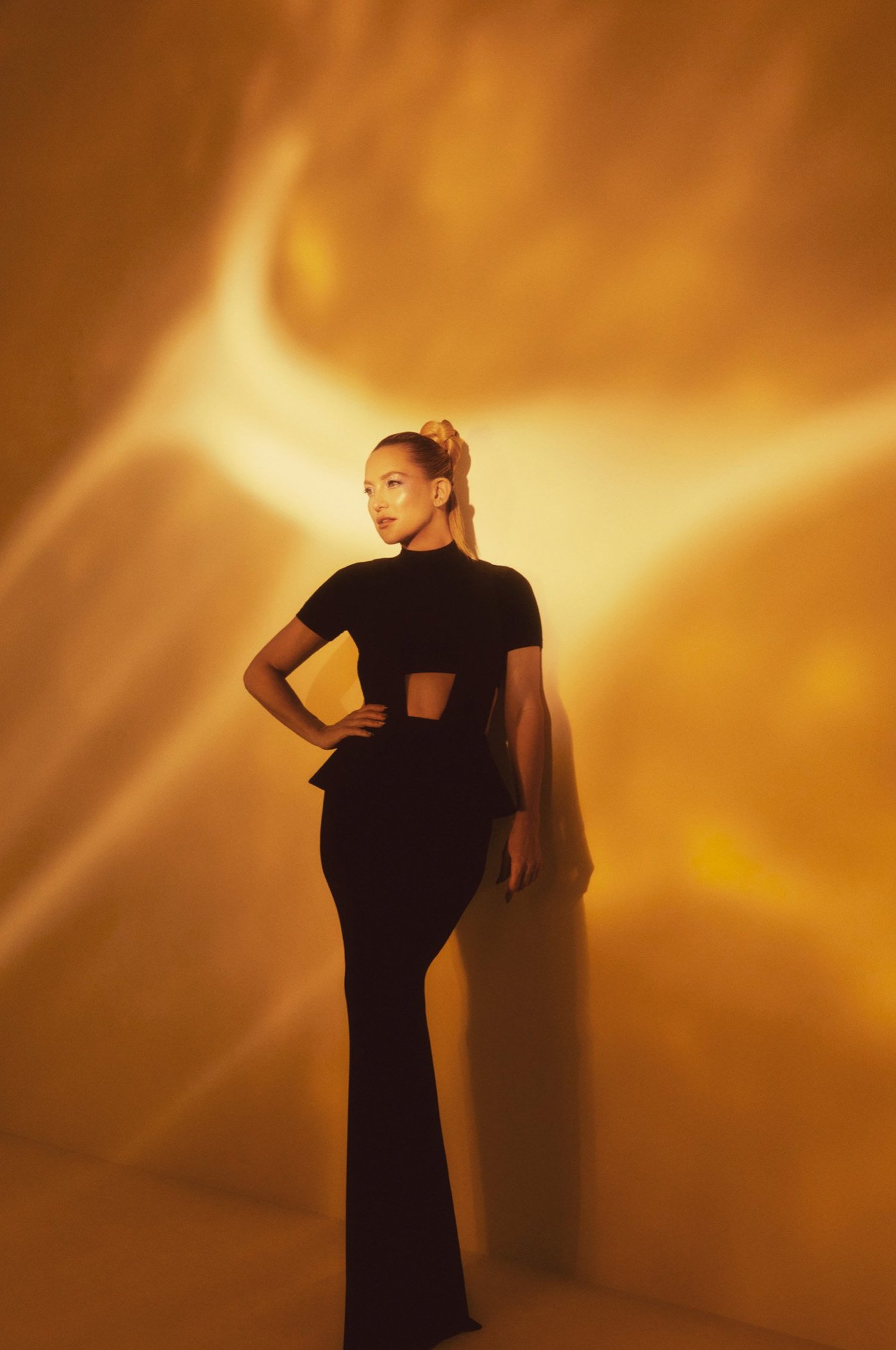 A woman in a black dress stands against a textured gold backdrop, captured in a medium shot with soft lighting.