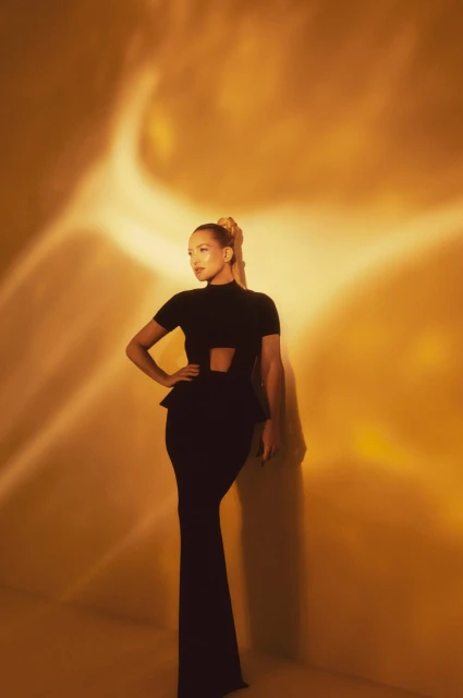 A woman in a black dress stands against a textured gold backdrop, captured in a medium shot with soft lighting.