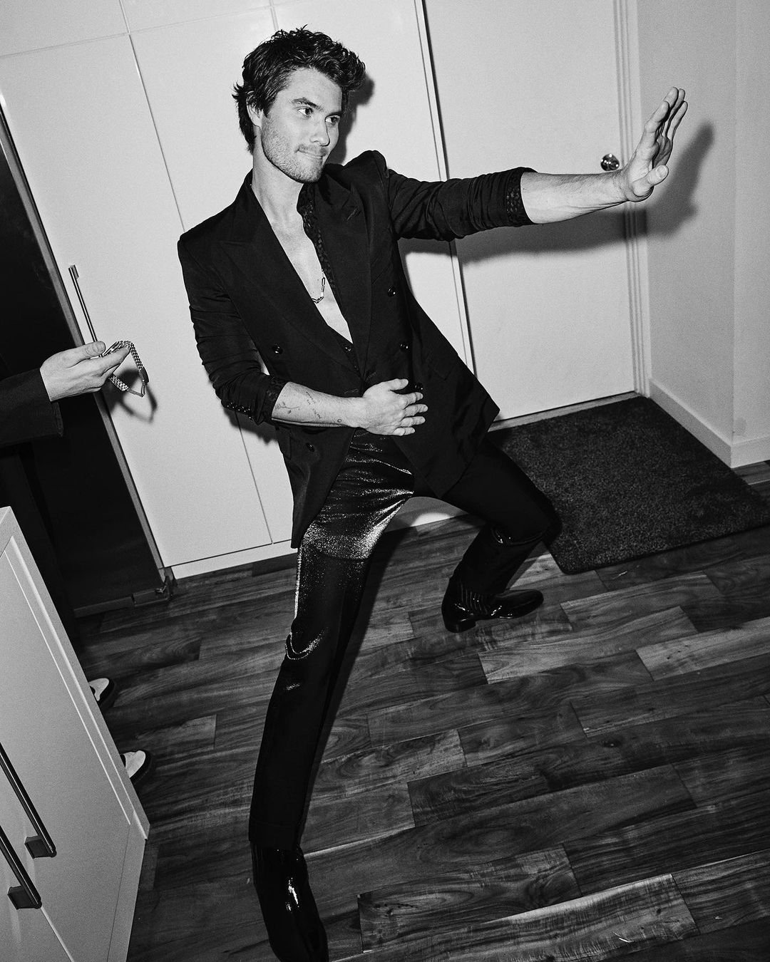 Black and white portrait of a man in a blazer and shiny pants posing indoors with an arm outstretched in a modern style.