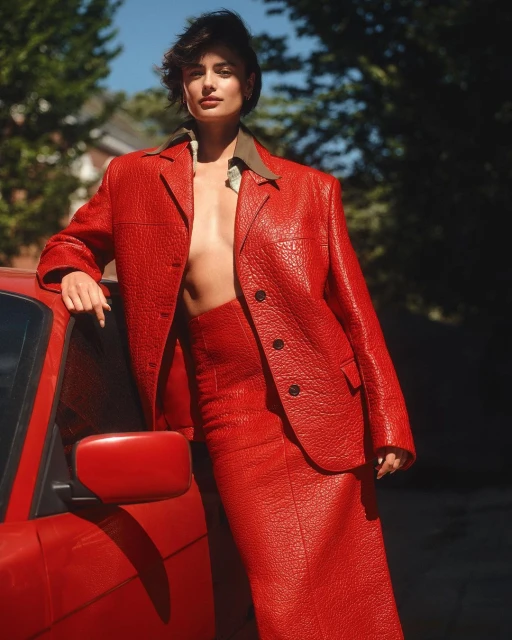 An adult female model poses with a red car, wearing a red blazer and skirt. The photo is shot outdoors on a sunny day.