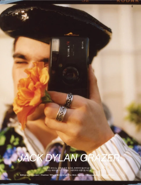 Portrait of Jack Dylan Grazer holding a camera and a flower in a close-up shot. He is wearing a beret and a patterned shirt.