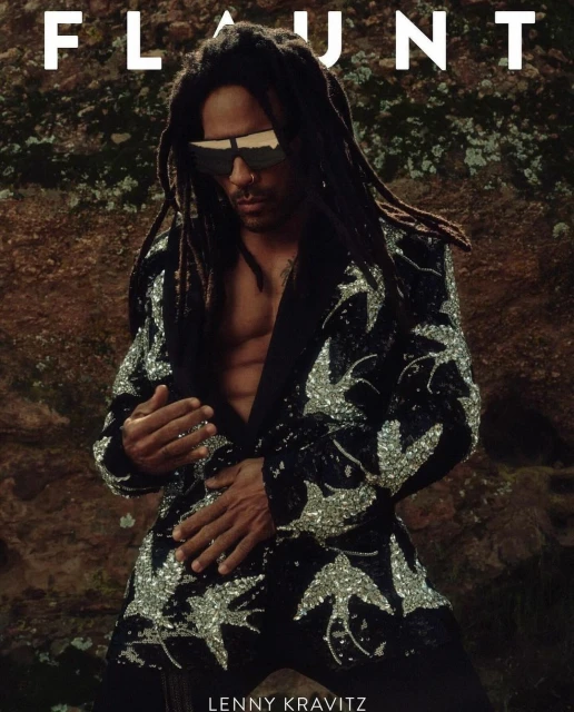 Lenny Kravitz poses in a black blazer with silver bird designs against a textured tan background for magazine cover art.