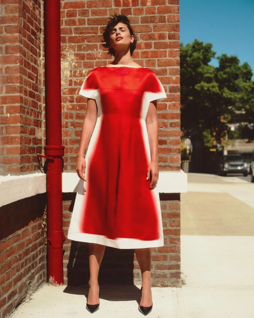 A woman in a striking red and white dress stands against a brick wall with natural light, showcasing fashion and urban style.