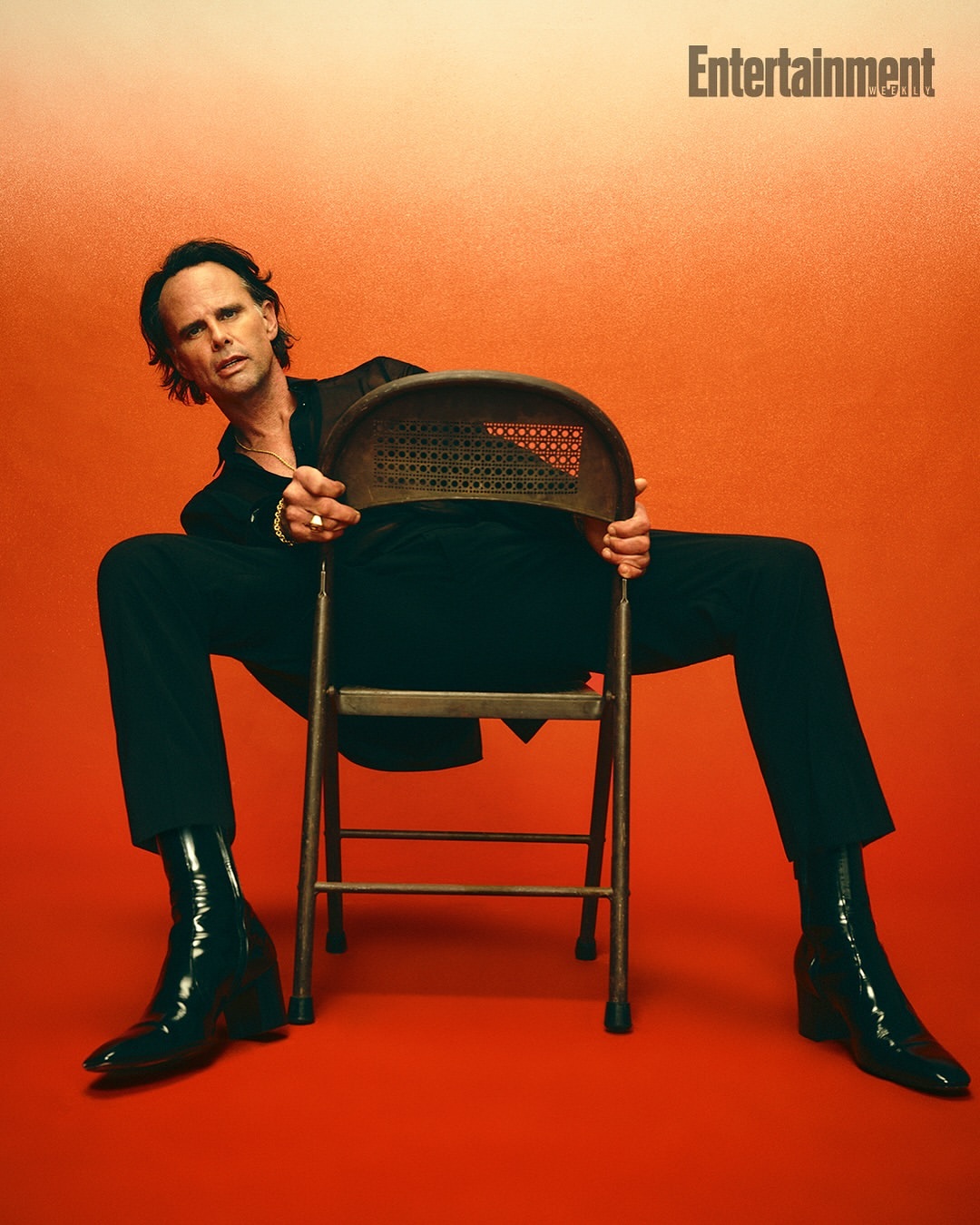 Adult male in black suit posing on metal chair with orange backdrop. Looking at the camera wearing black boots and wrist jewelry.