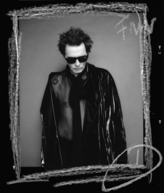 Black and white portrait of a mature adult man with sunglasses, wearing a vinyl jacket and scarf against a grey backdrop.