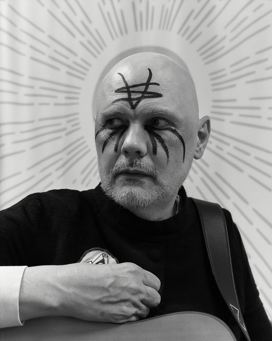 Black and white portrait of a bald adult male with face paint, wearing a dark sweater, and holding a guitar with graphic background.