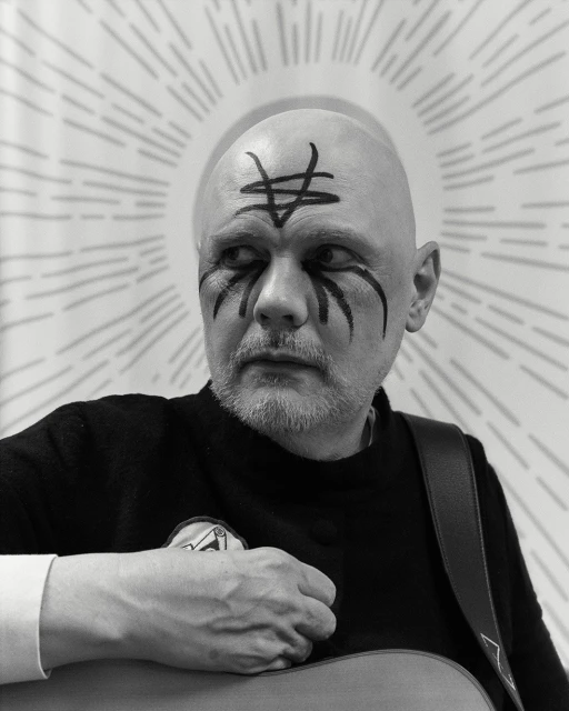 Black and white portrait of a bald adult male with face paint, wearing a dark sweater, and holding a guitar with graphic background.