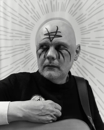 Black and white portrait of a bald adult male with face paint, wearing a dark sweater, and holding a guitar with graphic background.