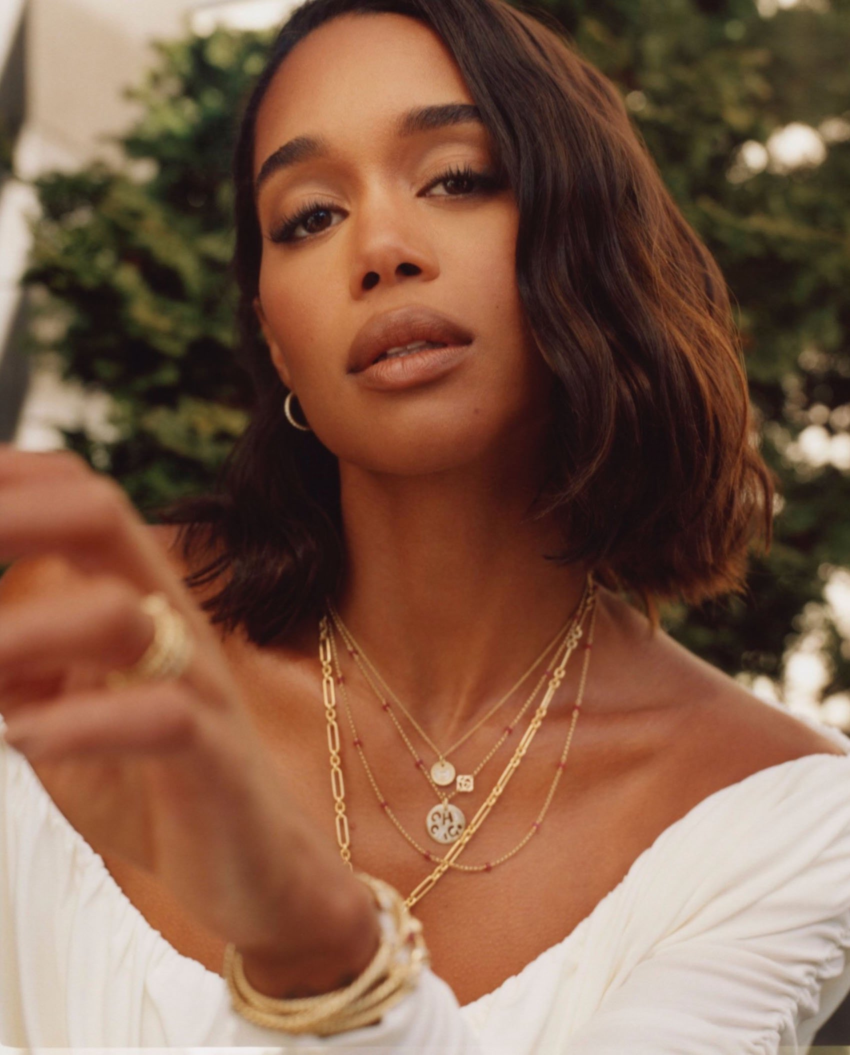 Close-up of woman with short dark hair wearing layered gold necklaces with white top and green leafy background. Beauty, fashion, portrait.