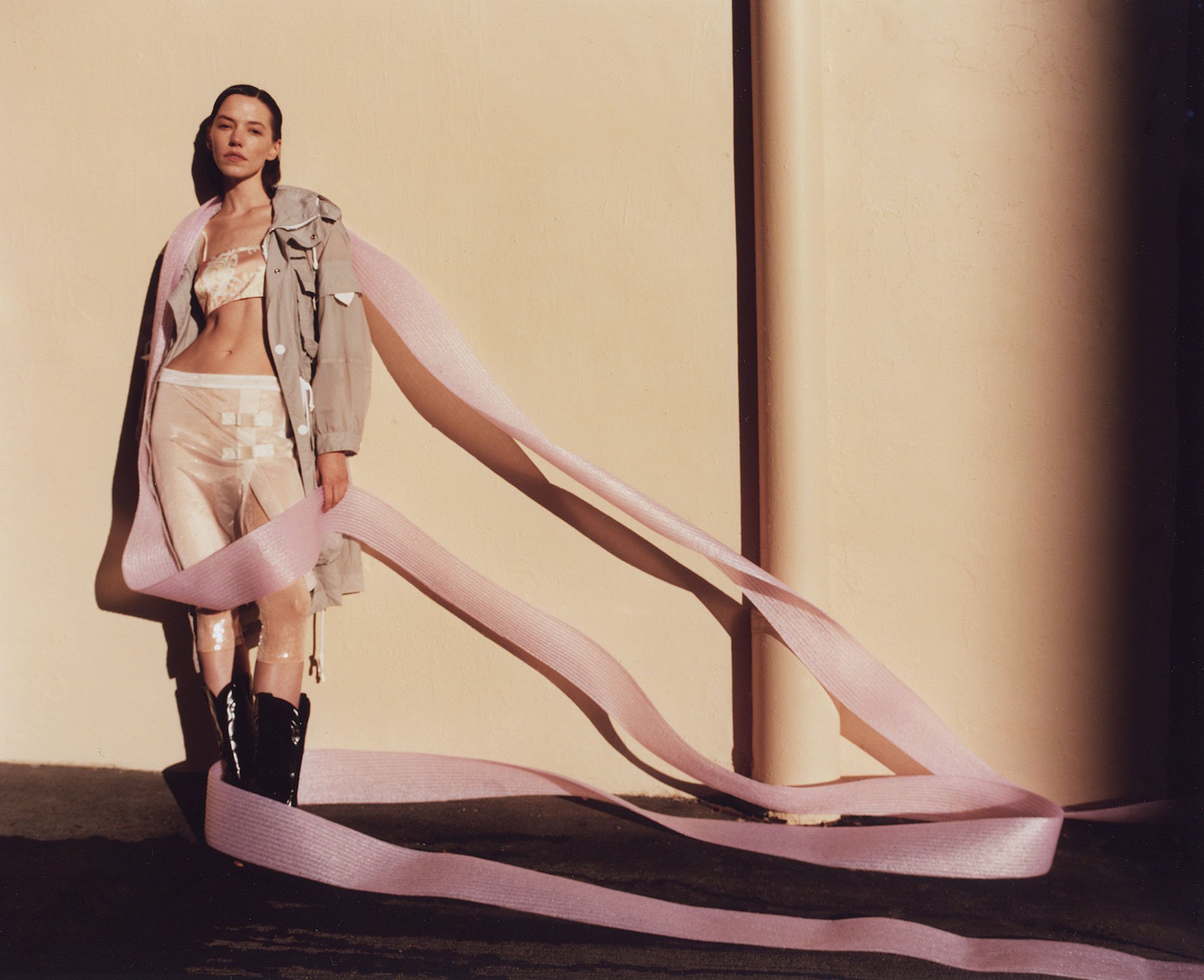 A woman in a light-colored outfit and black boots poses in front of a wall with a long pink ribbon wrapped around her in a studio setting.