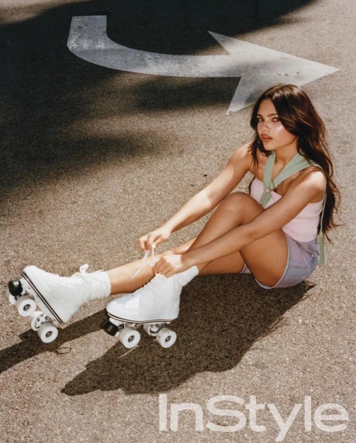Young woman with long brown hair sitting on the asphalt ground with roller skates ties her laces with an arrow on the ground behind.