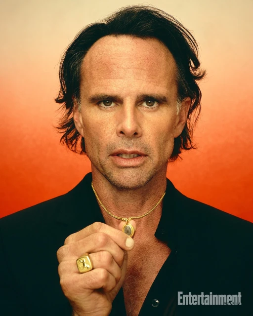 Portrait of an adult man with dark hair wearing a black shirt and gold jewelry against a gradient orange and white background.