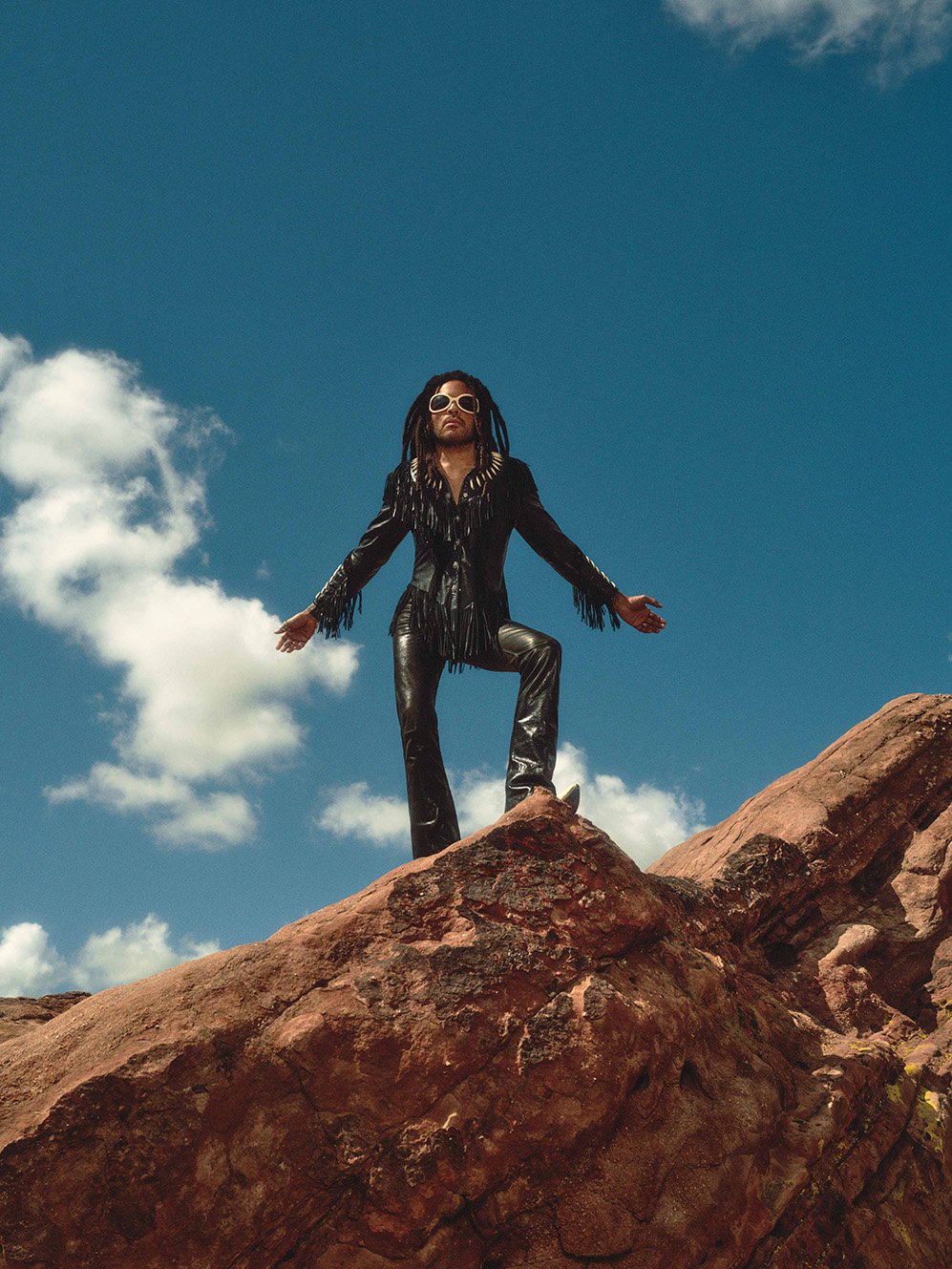Adult male with dreadlocks poses on a red rock formation against a blue sky wearing sunglasses, black jacket, and leather pants.