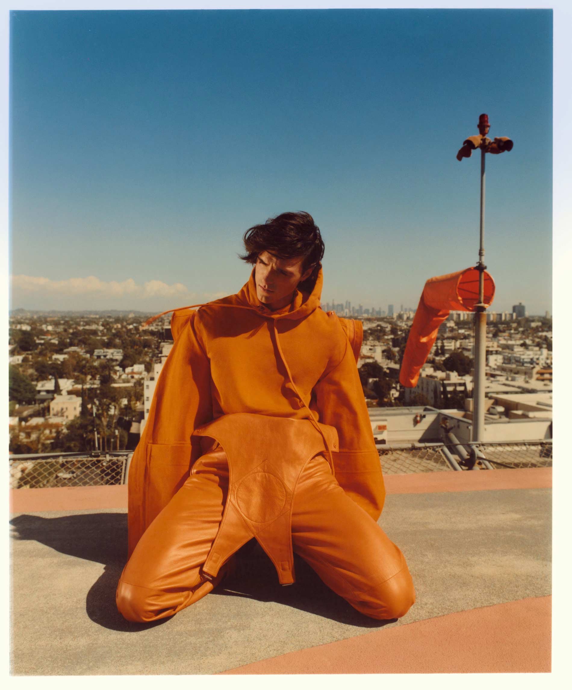 Adult male model in orange hooded costume kneeling on a rooftop with city skyline under a clear blue sky and orange windsock.
