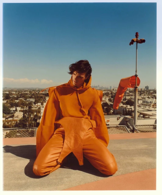Adult male model in orange hooded costume kneeling on a rooftop with city skyline under a clear blue sky and orange windsock.