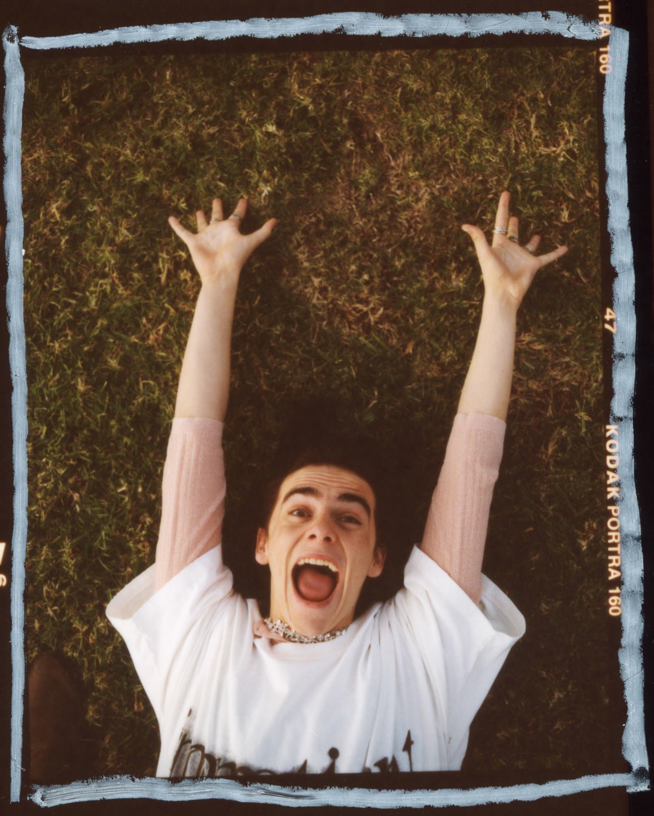 Young adult lies on grassy lawn with arms raised and mouth open, wearing a white graphic tee and beaded necklace, shot in daylight.