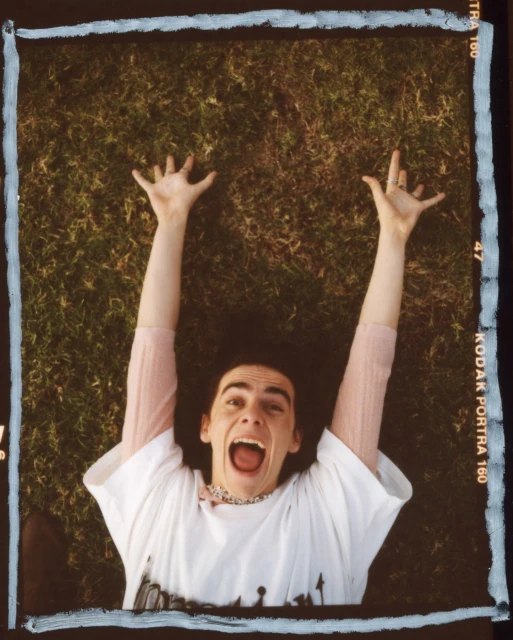 Young adult lies on grassy lawn with arms raised and mouth open, wearing a white graphic tee and beaded necklace, shot in daylight.